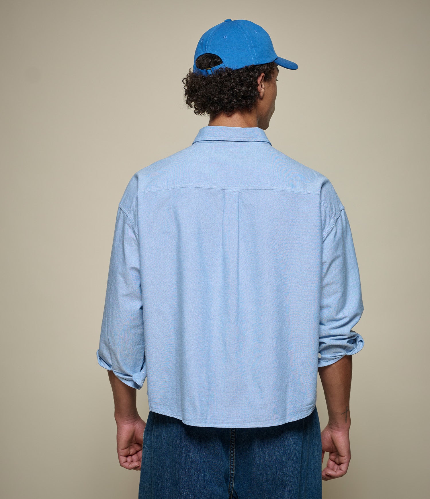 Man wearing a light blue cropped oxford shirt and blue cap from behind on a beige background