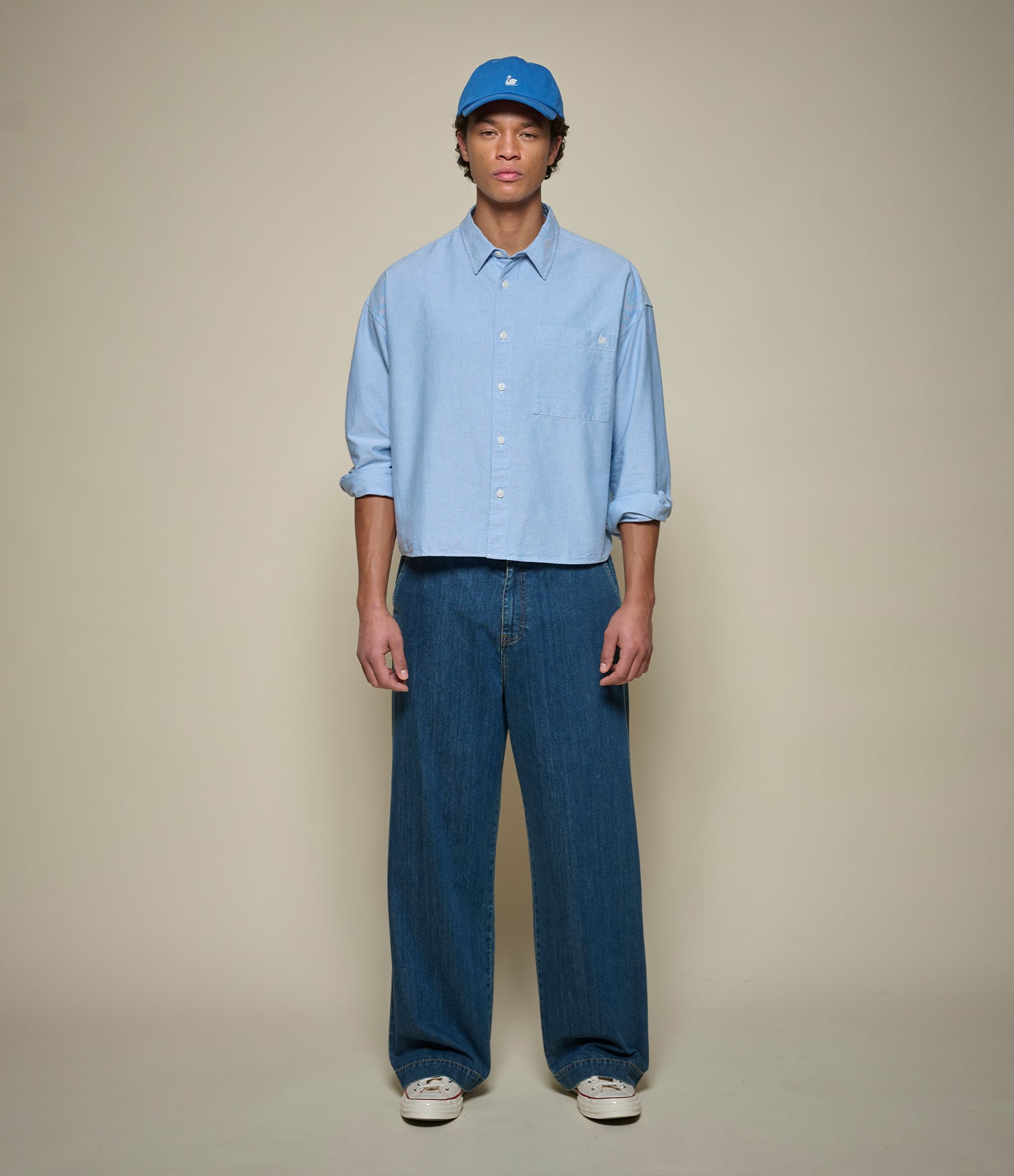 Man wearing a light blue cropped oxford shirt and dark blue jeans against a beige background