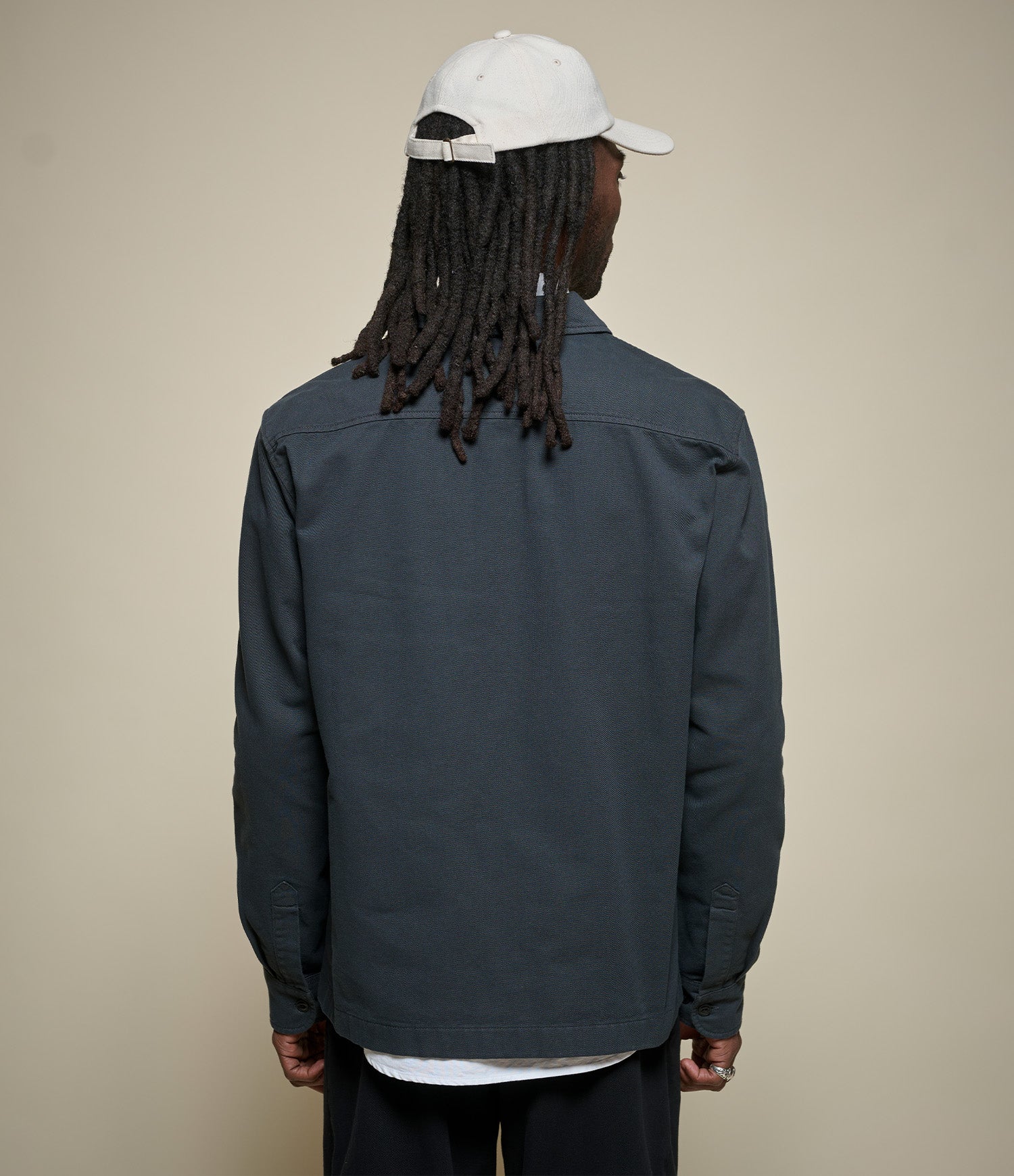 A man viewed from behind wearing a dark slate gray jacket, a white baseball cap, and black pants, standing against a solid beige background.