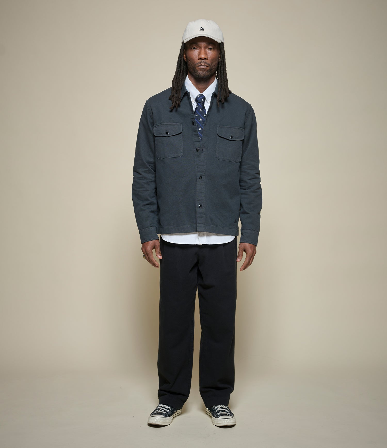 A man stands against a plain beige studio background wearing a dark slate blue button-up overshirt over a white collared shirt and patterned tie, with dark trousers, a white cap, and black-and-white sneakers.