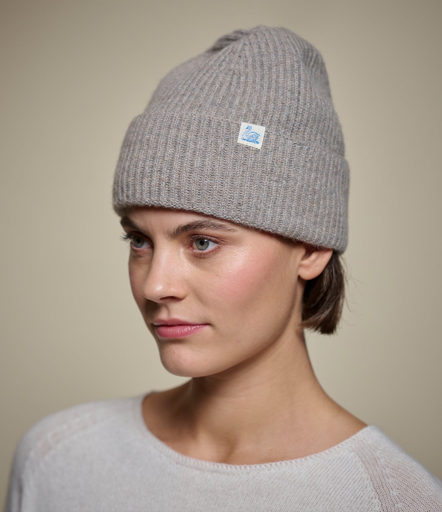 A woman wearing a light brown ribbed knit beanie hat with a small white brand tag on the side, photographed against a plain beige studio background.