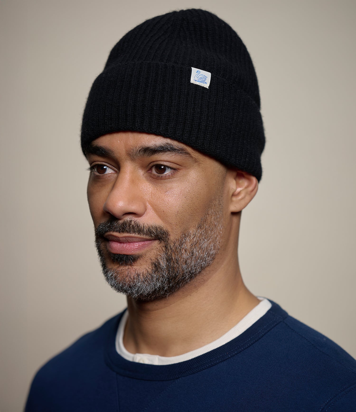 A man wearing a black ribbed knit beanie with a small swan logo patch on the side, photographed against a plain beige background.