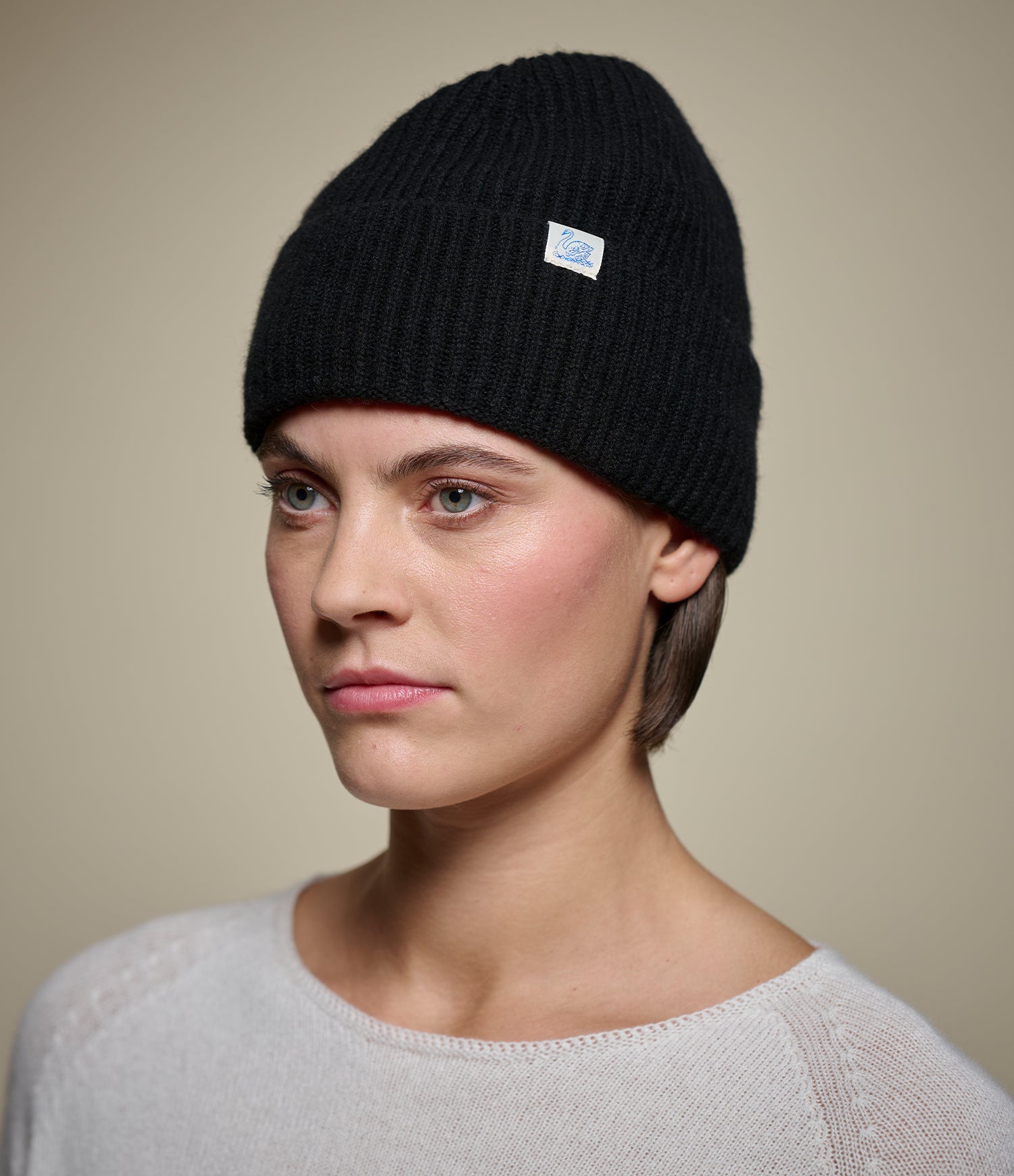Person wearing a black knit beanie with a swan logo on a beige background