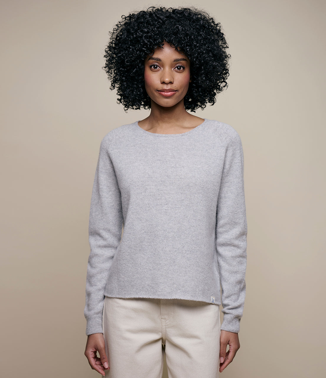 A person with curly hair wearing a long-sleeved, grey, round-neck Merino wool-cashmere-silk blend sweater paired with light-colored pants.