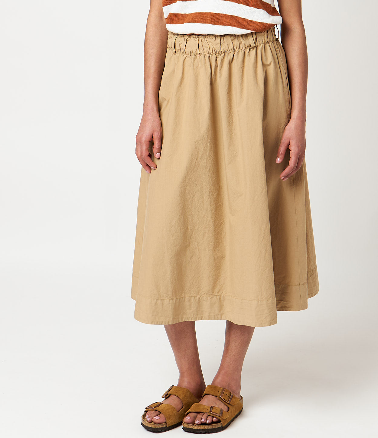 A person standing, wearing a beige knee-length skirt with a elastic waistband, paired with a striped top and brown sandals.