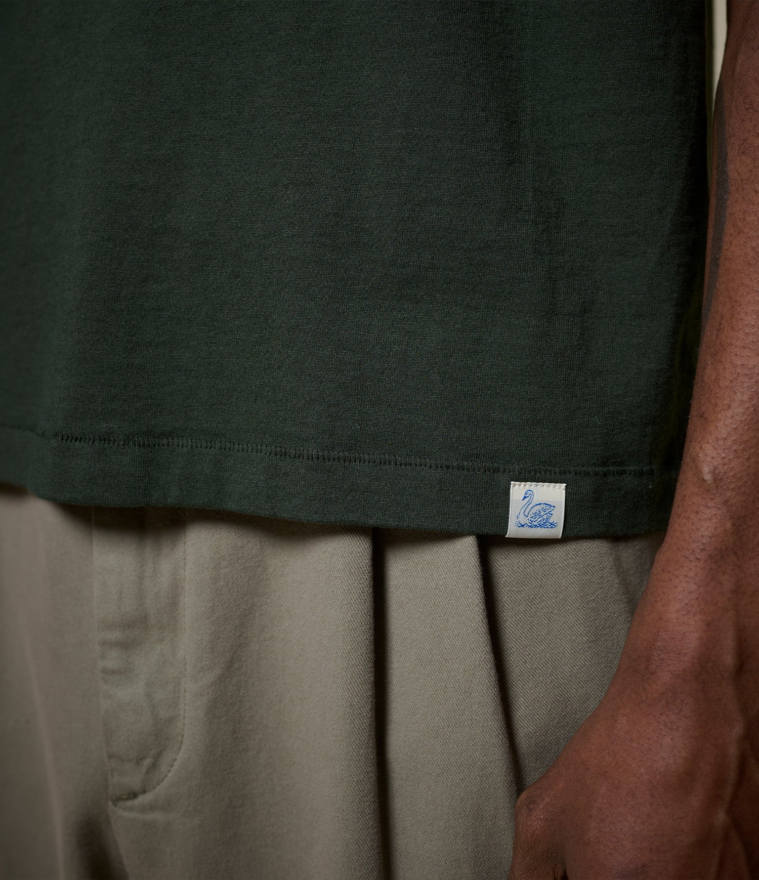 Close-up of a person wearing a dark green t-shirt with a visible Swan loop label on the hem.