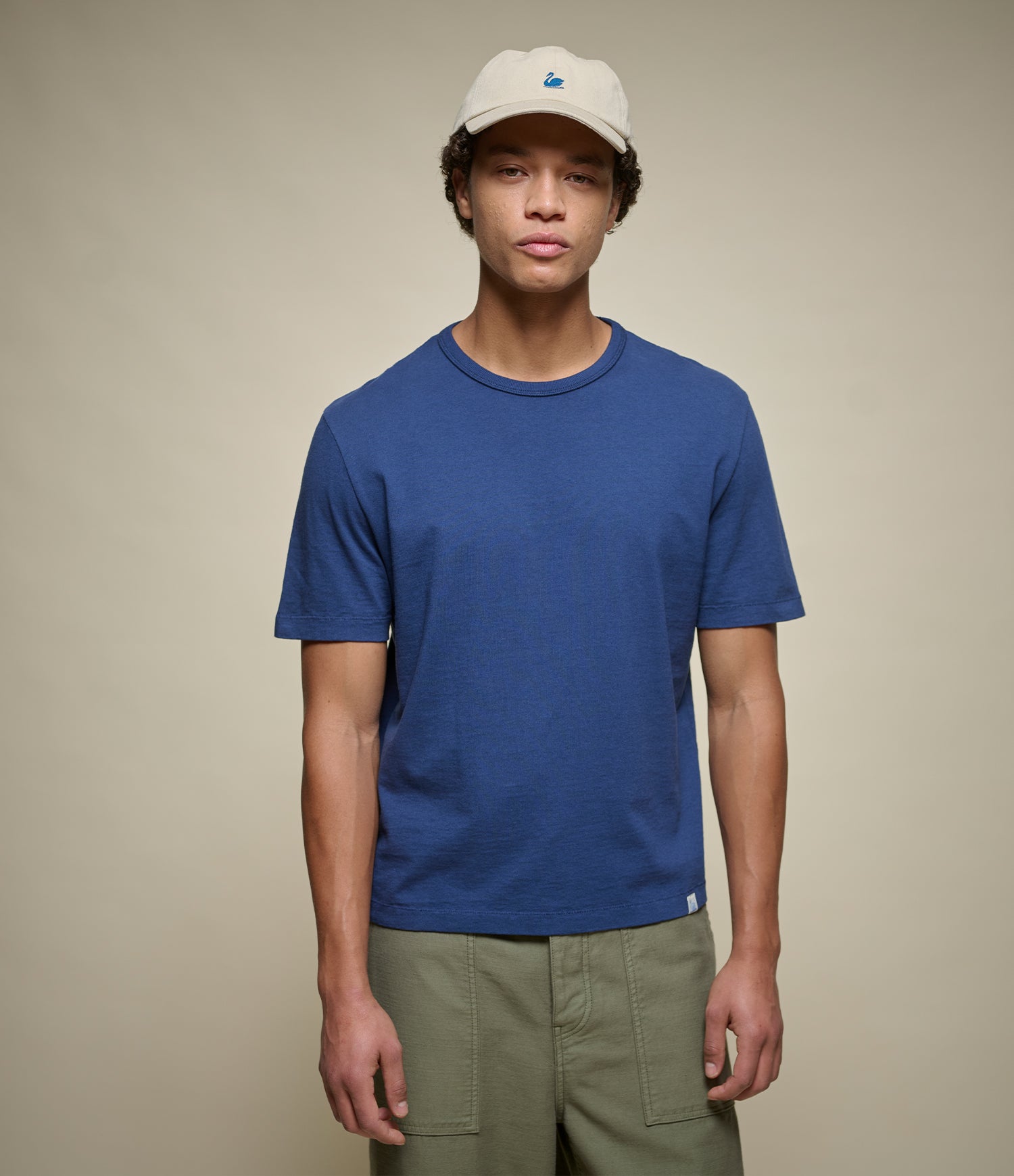 Person wearing a cropped blue crew-neck t-shirt and beige cap on a plain background