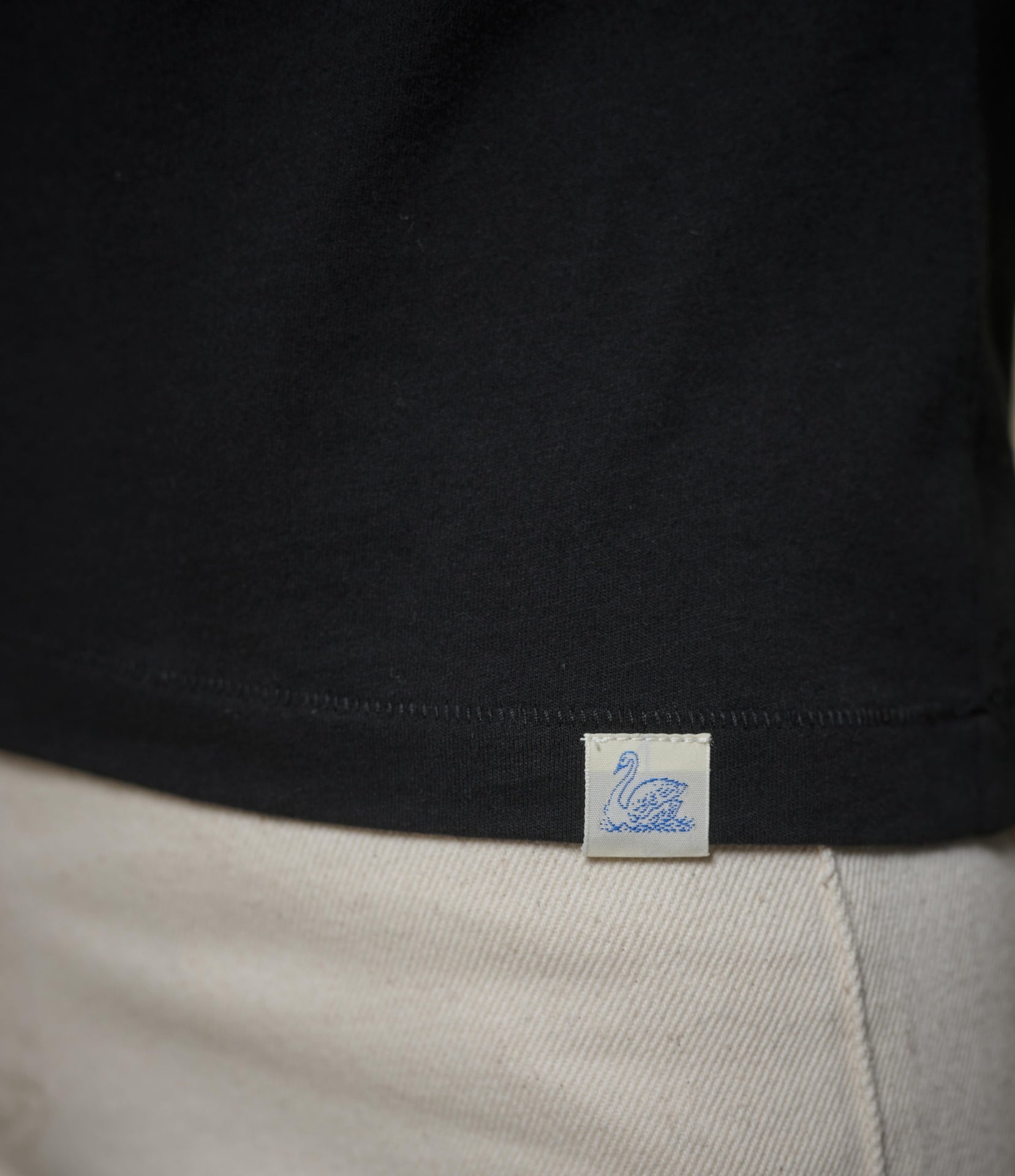 Close-up of a black t-shirt hem with a small white fabric brand tag featuring a blue swan logo, worn over light beige pants.