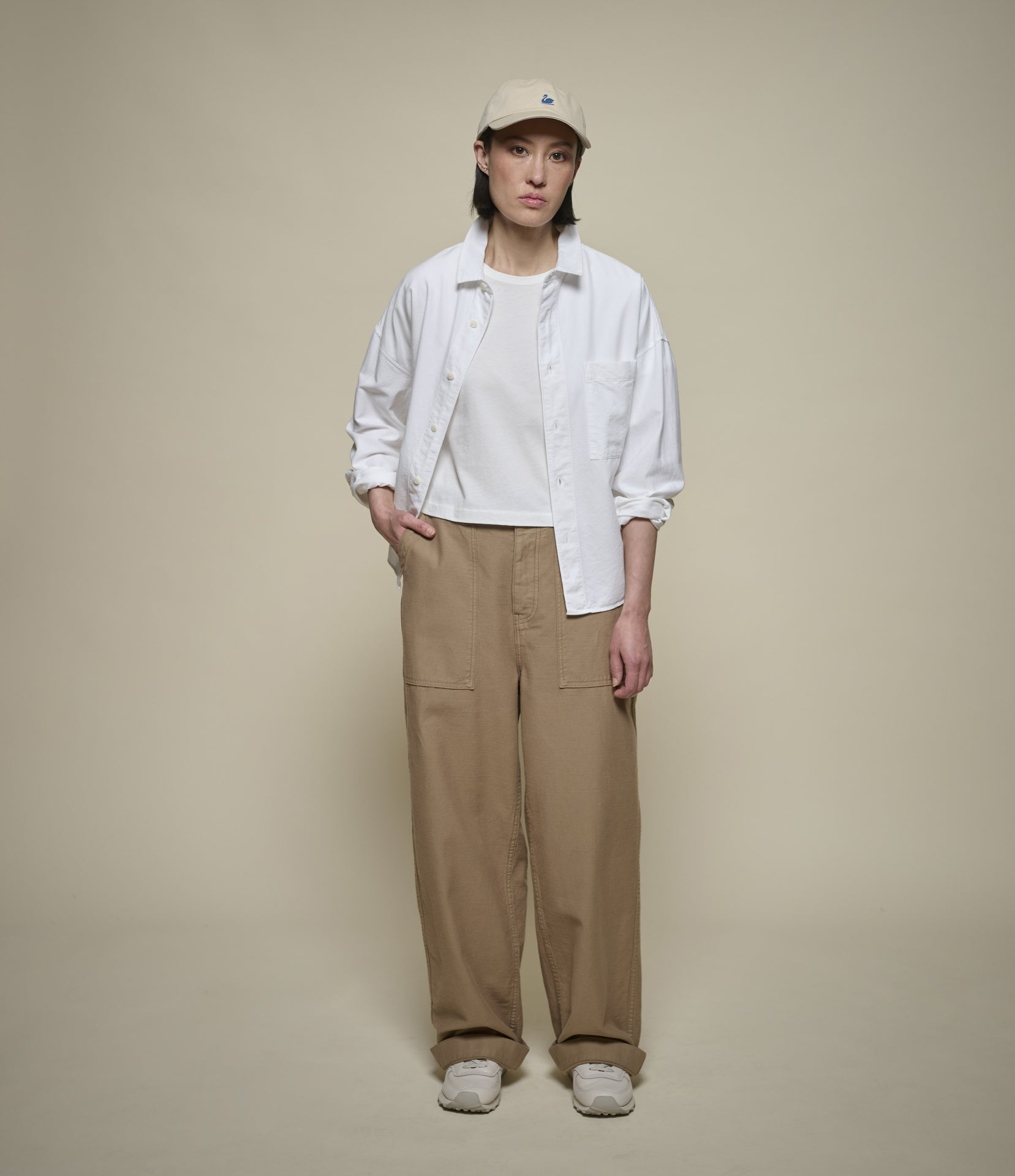 A model stands against a solid beige studio background wearing a white oxford shirt over a white t-shirt, beige wide-leg cargo pants, and a beige baseball cap.