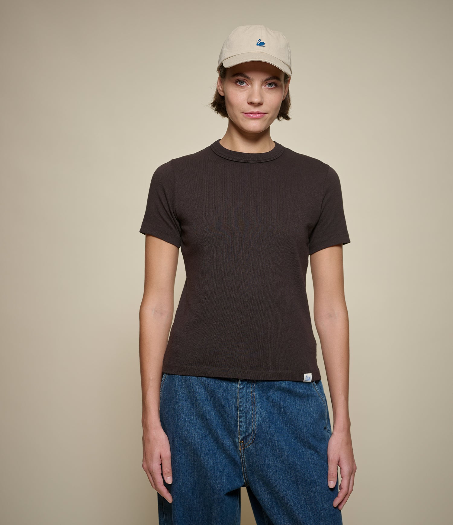 Woman wearing a dark brown short-sleeve crewneck t-shirt, blue jeans, and a beige baseball cap, standing against a solid beige background.