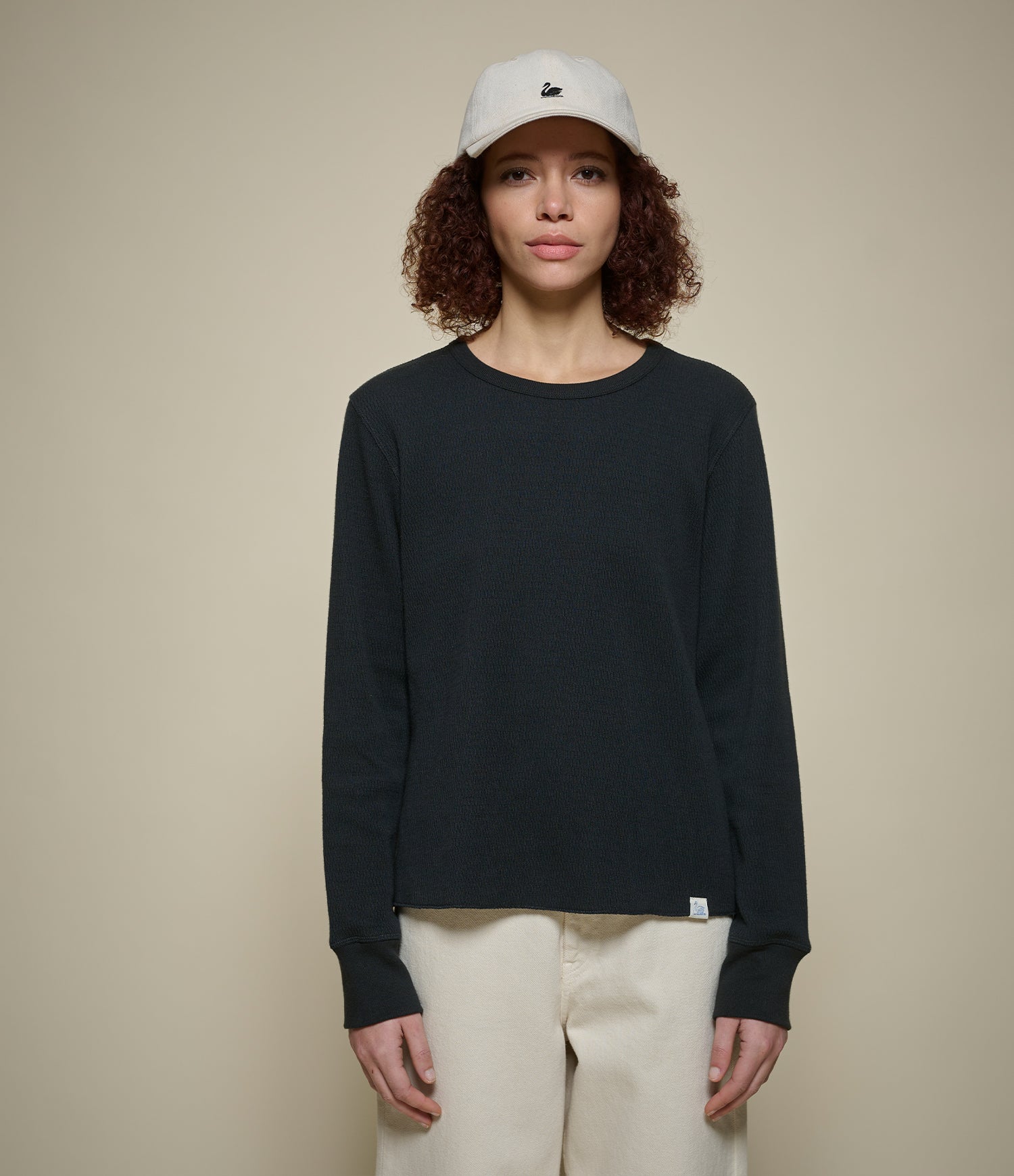 Person wearing a black long-sleeve thermal shirt, white pants, and a white cap with a logo on a beige background